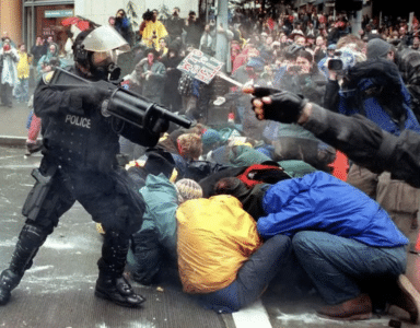 A police officer in riot gear and a gas mask aims a weapon at a group of protesters crouched on the ground, shielding themselves from pepper spray. The scene is chaotic, with other officers and a crowd of onlookers in the background, some holding cameras and signs. The image captures a moment of tension during a protest.